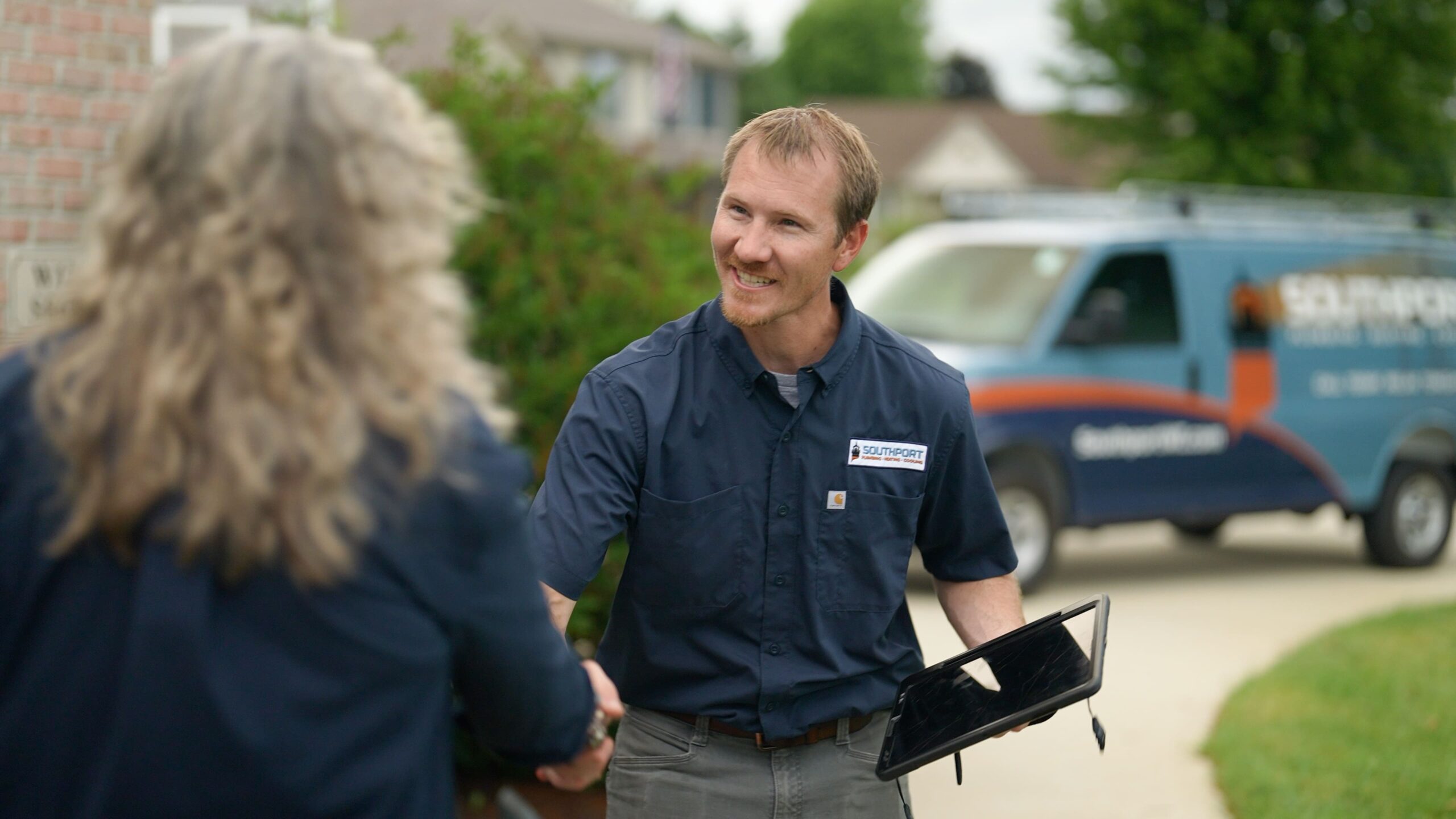 Man in a navy uniform shakes hands with a client outdoors, holding a clipboard while a blue service van sits in the background.