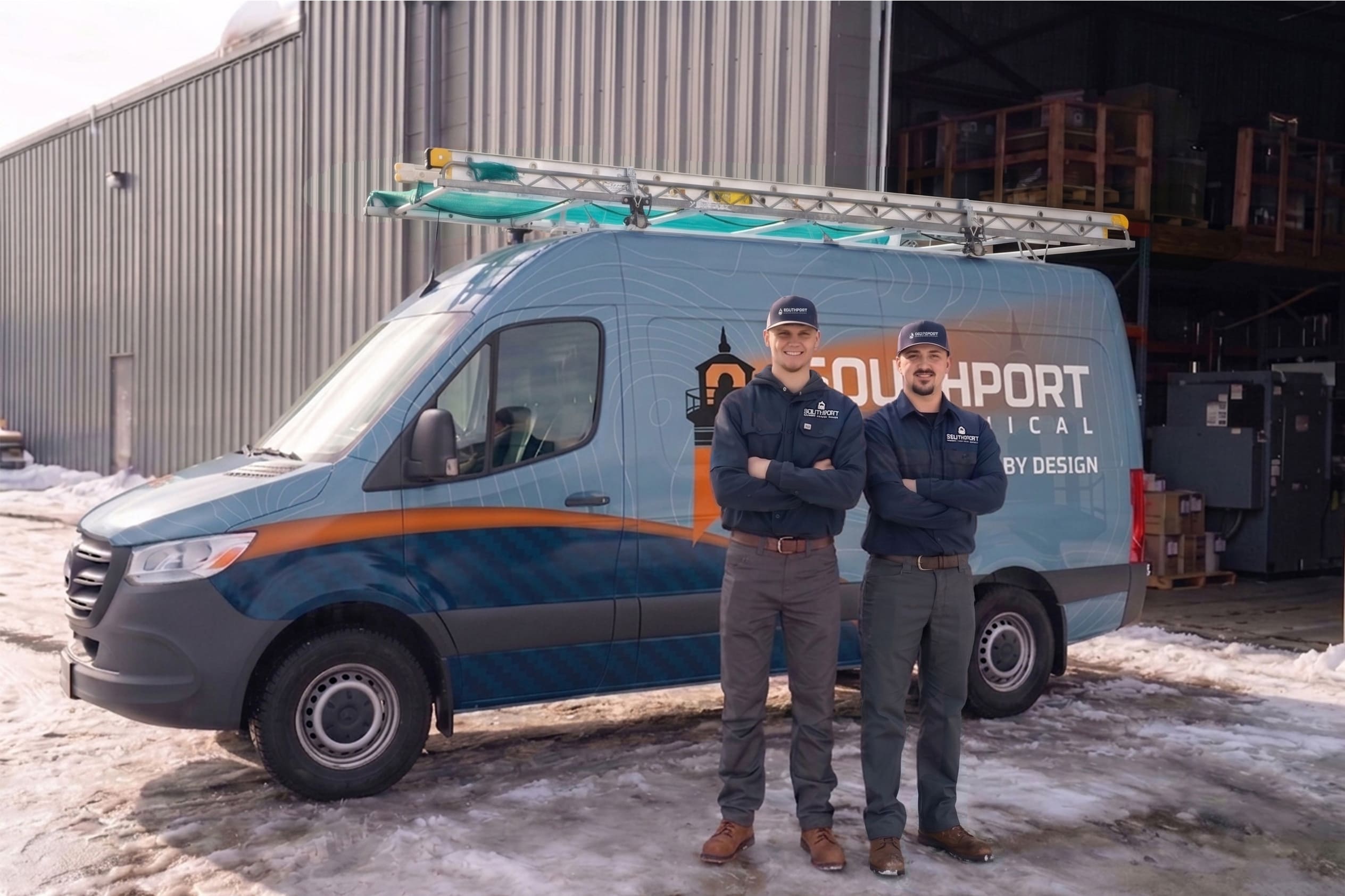Two workers in matching uniforms stand in front of a blue service van with a roof ladder rack in a snowy industrial yard.