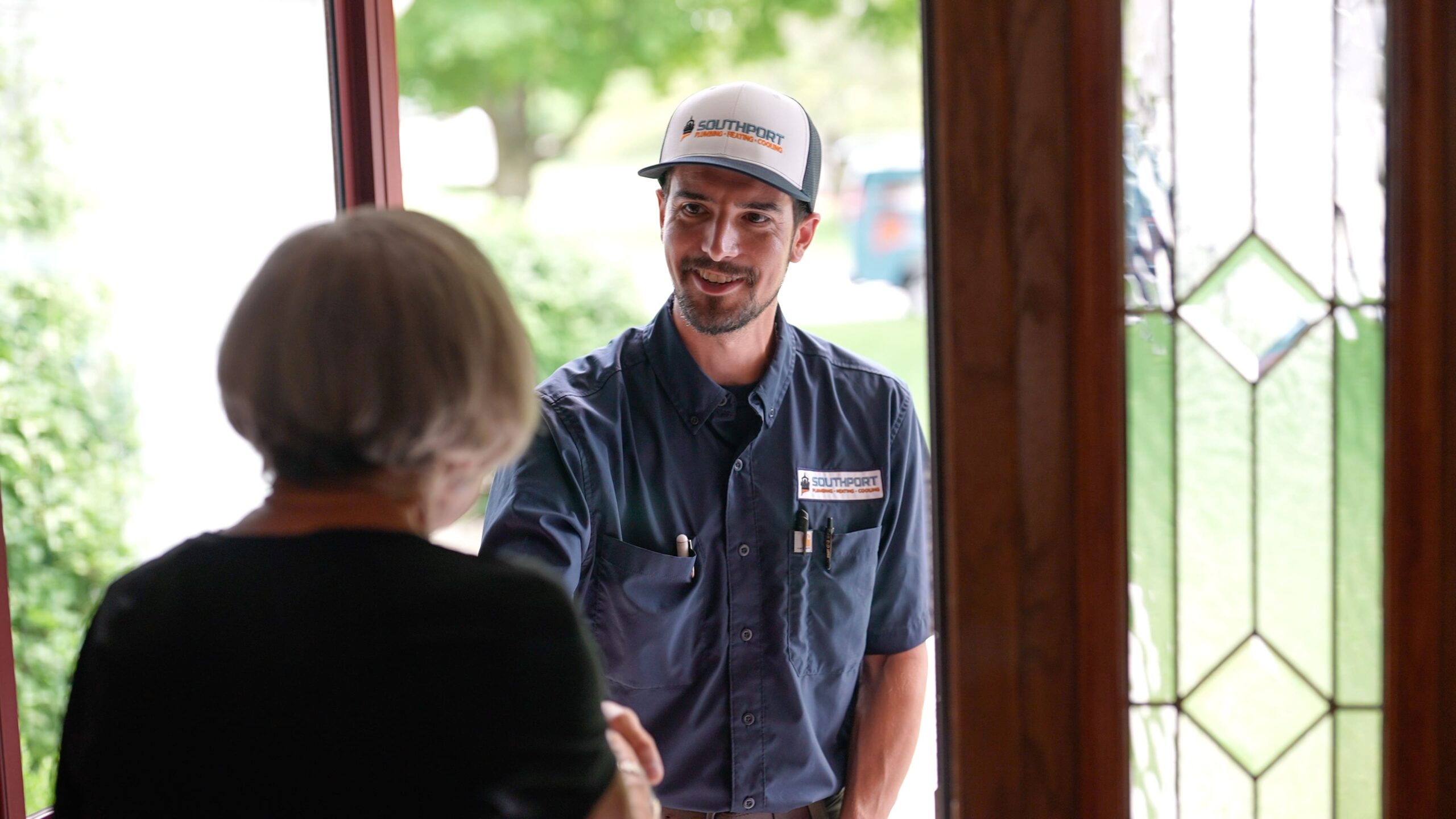 Smiling service worker in a blue uniform and white Southport cap greets a woman at a doorway.