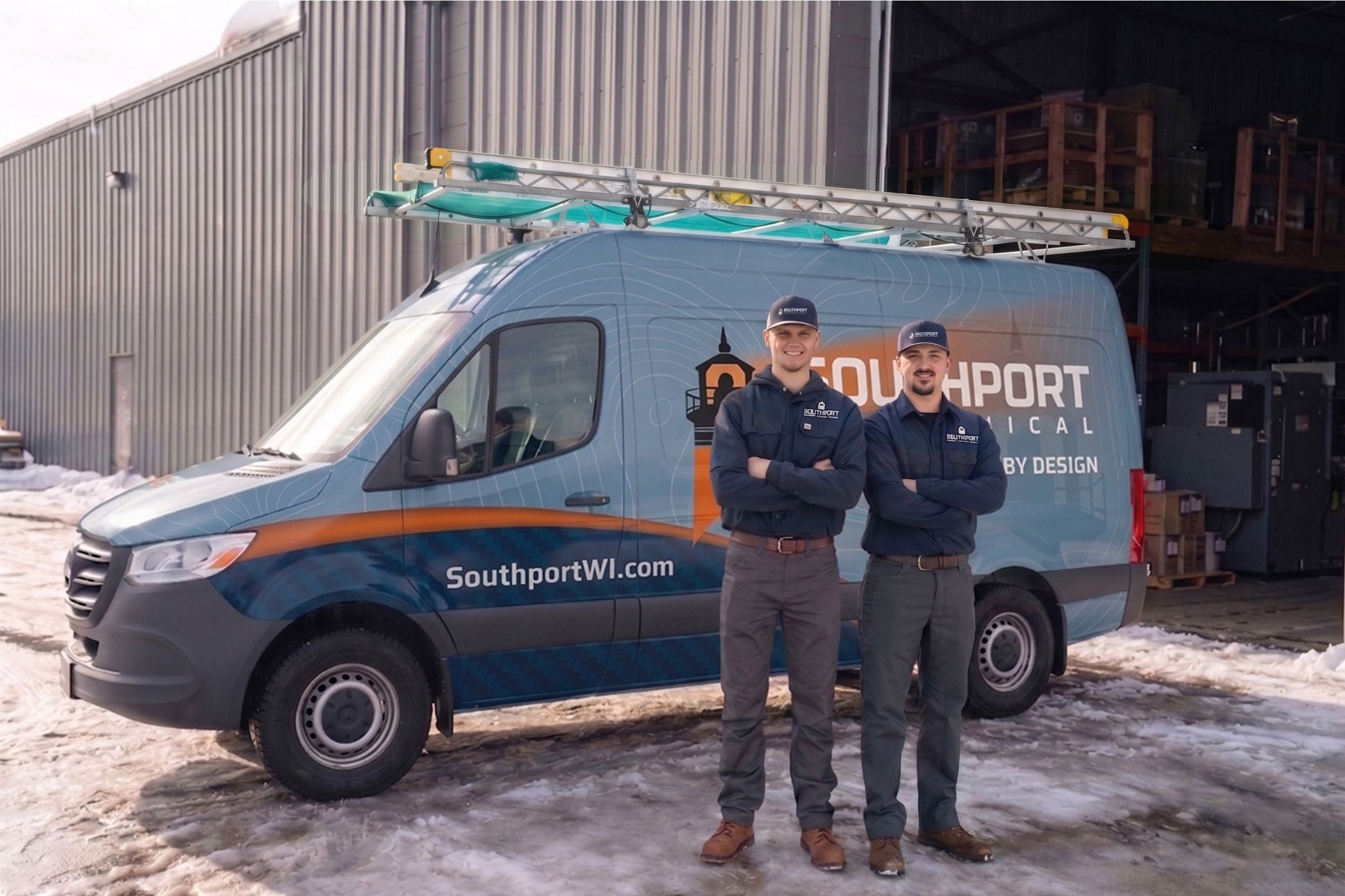 Two technicians in navy uniforms stand in a snowy lot beside a blue van with a roof ladder rack and warehouse in the background.