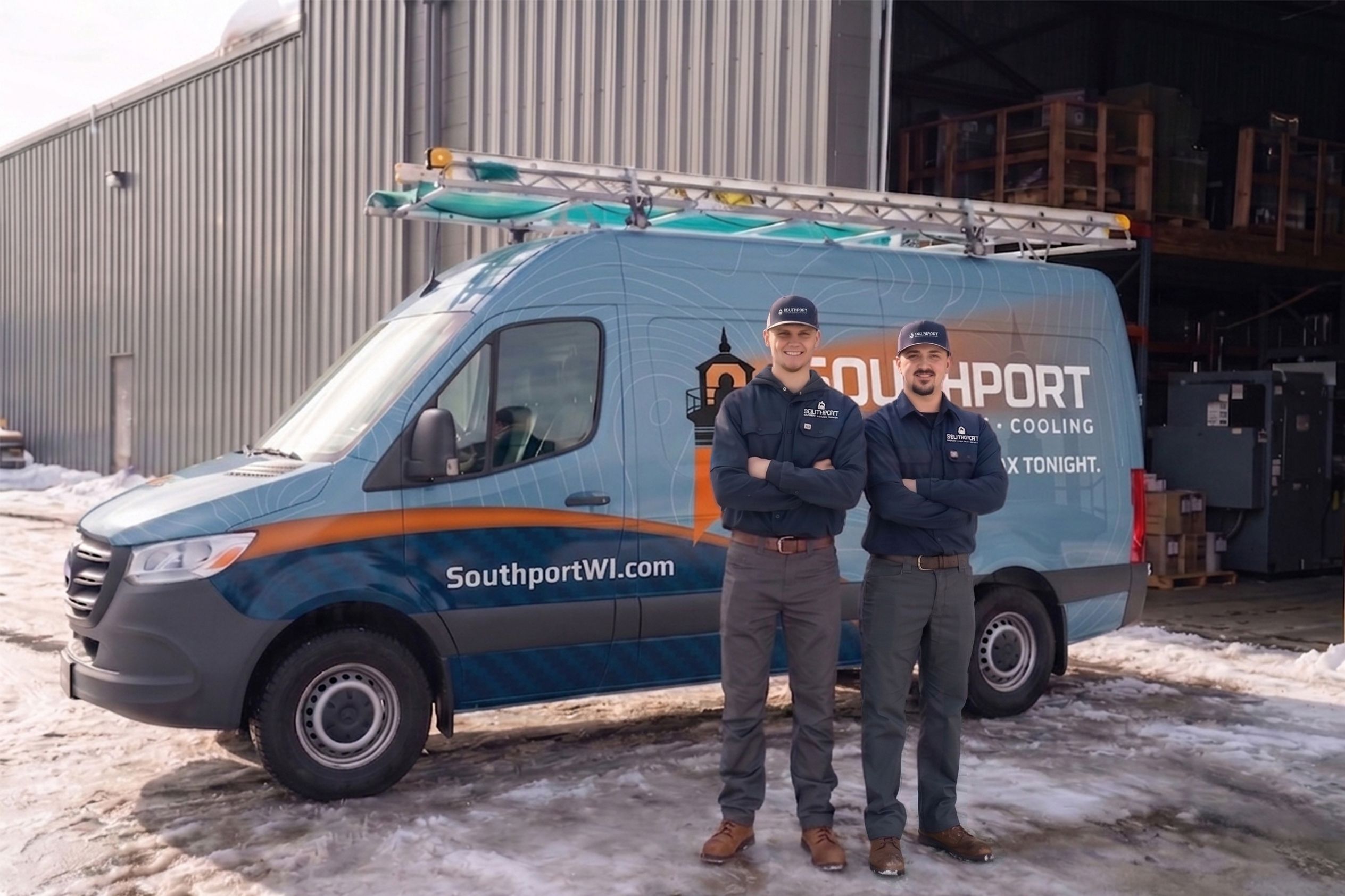 Two men in matching work uniforms pose with a blue service van outside a warehouse; ladder on roof and snow on the ground.