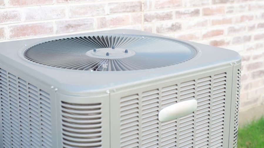 Outdoor air conditioner condenser unit with a circular top fan beside a brick wall.