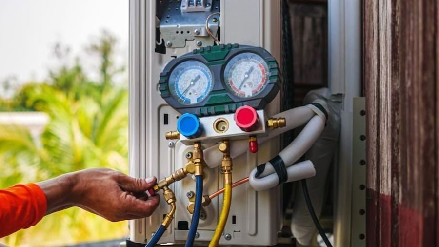 Technician connecting a blue and red manifold gauge set to an outdoor air conditioning unit with yellow refrigerant hoses attached