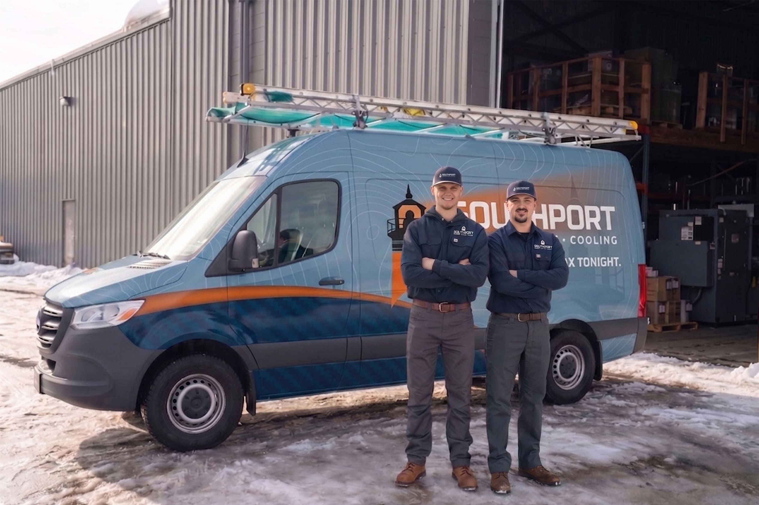 Two technicians in matching dark-blue uniforms stand with arms crossed in front of a blue service van with a roof ladder, outside a corrugated metal building in snow.