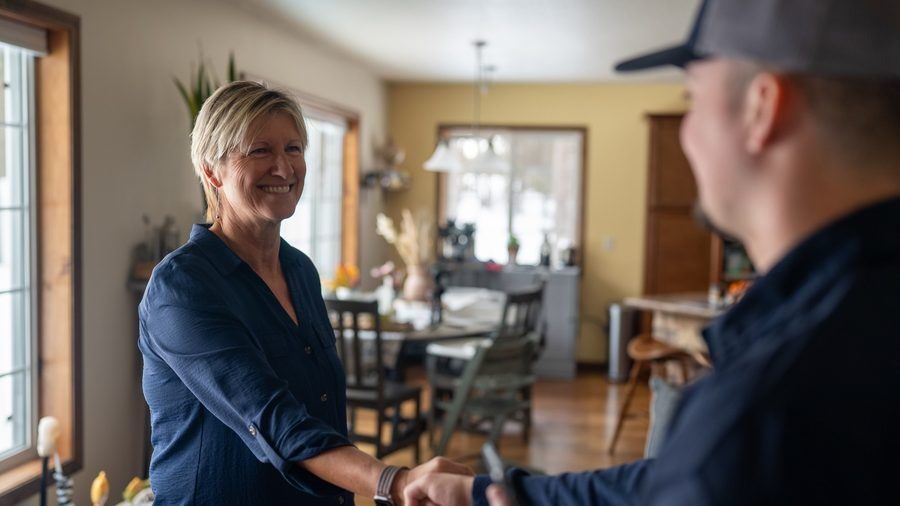 Smiling woman in a blue shirt shakes hands with a man in a cap in a bright, cozy kitchen/dining area at home.