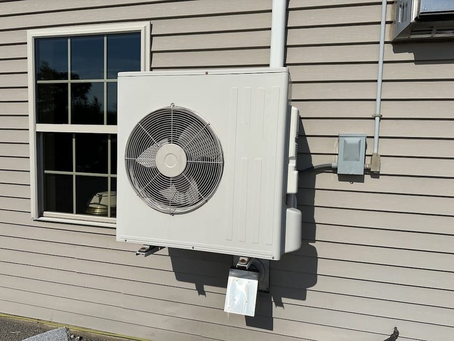 Outdoor HVAC unit mounted on the exterior wall beside a window, with electrical conduit and a metal support bracket.